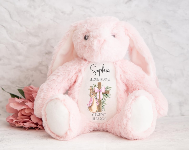 Pu&ograve; includere: Un coniglietto di peluche rosa con lunghe orecchie e zampe bianche. Il coniglietto ha un disegno stampato sul petto con una croce, un coniglio e il nome "Sophia" con la data "11.08.2024".