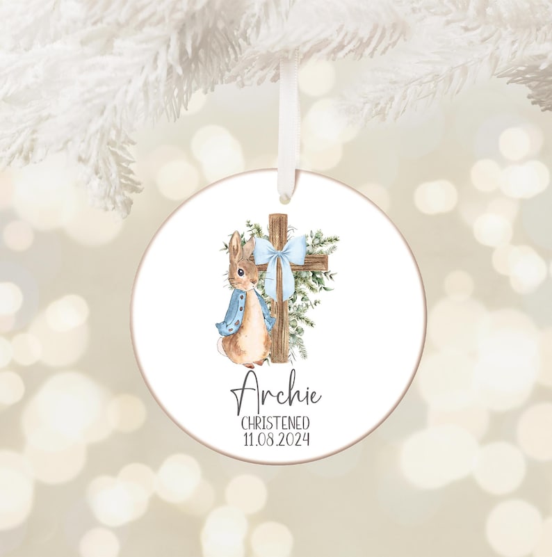 Pu&ograve; includere: Ornamento natalizio rotondo e bianco con un'illustrazione ad acquerello di un coniglietto con una giacca blu accanto a una croce di legno adornata da un fiocco blu e verde. Il nome "Archie" e la data "11.08.2024" sono stampati sotto.