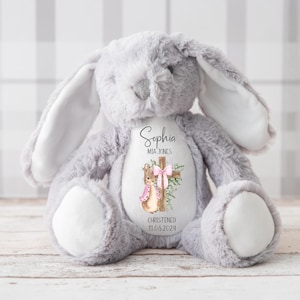 Pu&ograve; includere: Un coniglietto di peluche grigio con dettagli bianchi sulle orecchie e sulle zampe. Il coniglietto ha un disegno stampato sul petto con una croce, un coniglietto e il nome "Sophia Mia Jones" con la data "11.06.2024".