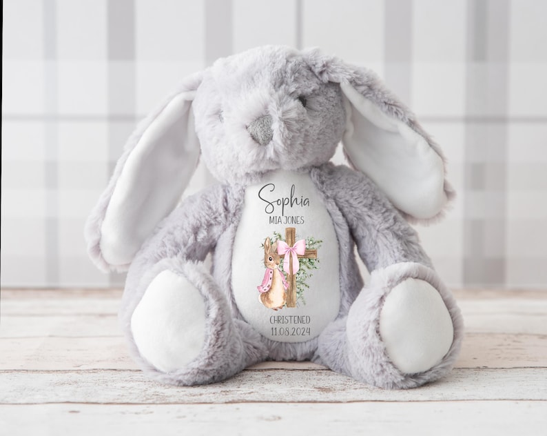 Pu&ograve; includere: Un coniglietto di peluche grigio con dettagli bianchi sulle orecchie e sulle zampe. Il coniglietto ha un disegno stampato sul petto con una croce, un coniglietto e il nome "Sophia Mia Jones" con la data "11.06.2024".