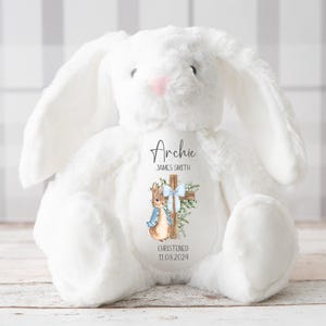 Pu&ograve; includere: Un coniglietto di peluche bianco con lunghe orecchie e naso rosa. Il coniglietto ha una toppa decorativa con il nome "Archie", una croce, un'illustrazione di coniglio e la data "11.08.2024". Il coniglietto &egrave; su una superficie chiara.