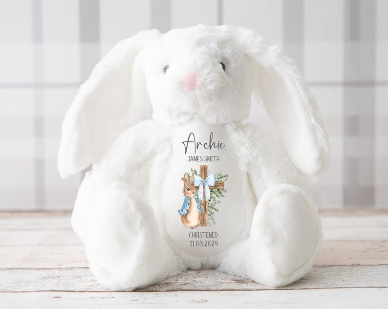 Pu&ograve; includere: Un coniglietto di peluche bianco con lunghe orecchie e naso rosa. Il coniglietto ha una toppa decorativa con il nome "Archie", una croce, un'illustrazione di coniglio e la data "11.08.2024". Il coniglietto &egrave; su una superficie chiara.