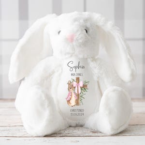 Pu&ograve; includere: Un coniglietto di peluche bianco con lunghe orecchie e un naso rosa. Il coniglietto ha un disegno stampato con una croce, un coniglietto con un vestito rosa, il nome "Sophia", "Mia Jones" e la data "11.08.2024".