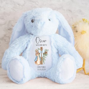 Pu&ograve; includere: Un coniglio di peluche azzurro con lunghe orecchie e pancia bianca. Il coniglio ha un disegno stampato di una croce, un coniglio e il nome "Oscar" con la data "11.08.2024". Un pulcino giallo &egrave; sulla destra.