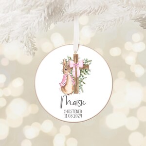 Pu&ograve; includere: Ornamento rotondo bianco con un'illustrazione ad acquerello di un coniglietto con una sciarpa rosa e una croce con un fiocco rosa. Il nome "Maisie" e la data "11.08.2024" sono stampati sotto. Un nastro bianco &egrave; attaccato per appenderlo.