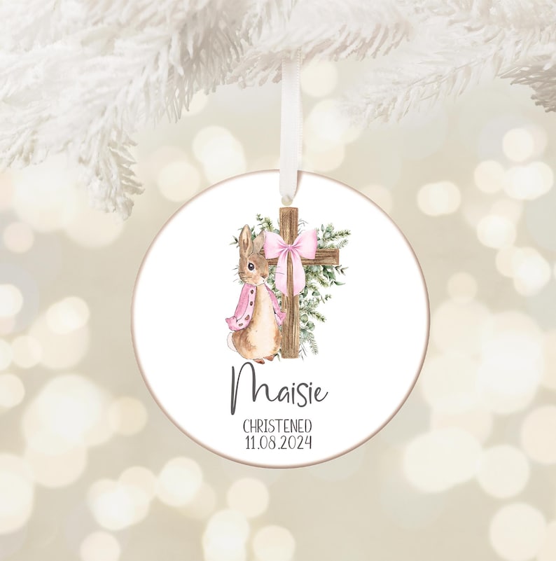 Pu&ograve; includere: Ornamento rotondo bianco con un'illustrazione ad acquerello di un coniglietto con una sciarpa rosa e una croce con un fiocco rosa. Il nome "Maisie" e la data "11.08.2024" sono stampati sotto. Un nastro bianco &egrave; attaccato per appenderlo.