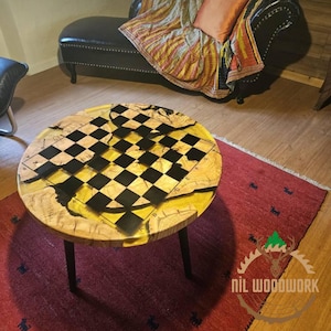 Puede incluir: Una mesa redonda de madera con un diseño de tablero de ajedrez en blanco y negro incrustado en la superficie. La mesa tiene patas de metal negro y está sobre una alfombra roja. El texto "NIL WOODWORK" es visible en la esquina inferior derecha de la imagen.