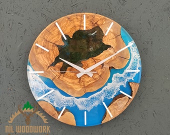 Può includere: Un orologio rotondo in legno con un'intarsio in resina epossidica blu e bianca. Il quadrante dell'orologio presenta indici orari bianchi e lancette bianche. L'intarsio in resina ricorda un'onda che si infrange su una costa.