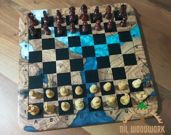 May include: A wooden chessboard with a blue and black epoxy resin inlay. The board has a light wood finish and features black and white squares. The chess pieces are made of wood and are arranged on the board in a starting position. The board is labeled with numbers and letters along the edges.