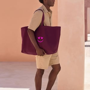 Puede incluir: Un bolso tote grande, de color burdeos, con un diseño de carita sonriente rosa. El bolso lo lleva una persona que viste camisa y pantalones cortos color beige. El bolso tote tiene un diseño sencillo y está hecho de un material resistente.