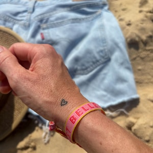 Könnte beinhalten: Ein Handgelenk mit einem rosa und gelben Armband, auf dem in rosa Buchstaben "I Believe" steht. Die Person hat ein kleines Herz-Tattoo am Handgelenk.