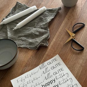 Könnte beinhalten: Ein weißes Blatt Papier mit dem Text "Fifty but fabulous Alles GUTE", der dreimal wiederholt wird. Unter dem Text werden die Worte "happy birthday" viermal in einer handschriftlichen Schrift wiederholt.