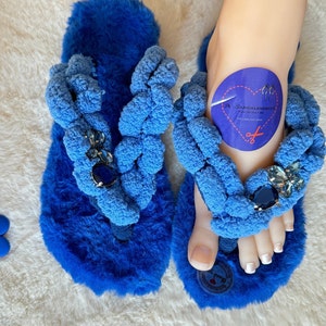 May include: Blue fuzzy slippers with a braided strap and jeweled embellishments. The slippers have a heart-shaped tag with the text "SANDALENDELLX - Feel for the love - For Life and Style".