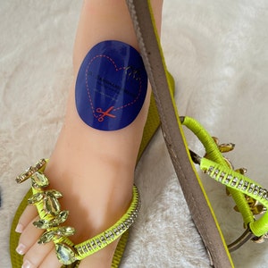 May include: Lime green flip flops with a rhinestone strap and gold-tone embellishments. The flip flops have a textured sole and are decorated with a blue circle sticker that says "SANDALEN PARADISE" with a heart and scissors.