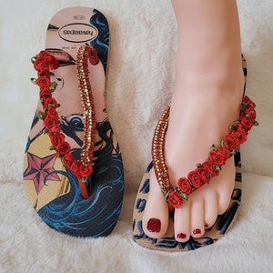 May include: A pair of flip-flops with a Wonder Woman graphic and red flower embellishments. The flip-flops are made by the brand "SPRINKBABY".