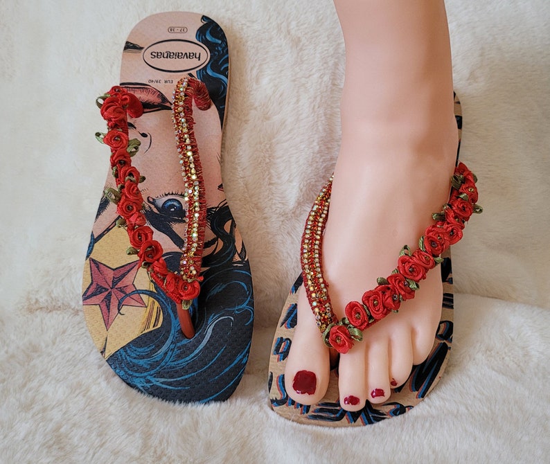May include: A pair of flip-flops with a Wonder Woman graphic and red flower embellishments. The flip-flops are made by the brand "SPRINKBABY".
