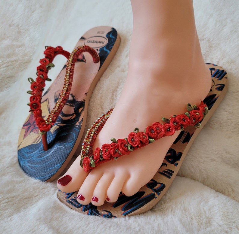 May include: A pair of brown flip-flops with a Wonder Woman graphic and red flower embellishments.