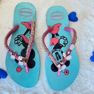 May include: A pair of blue flip-flops with pink straps and rhinestones. The flip-flops feature a cartoon image of Mickey Mouse and Minnie Mouse.