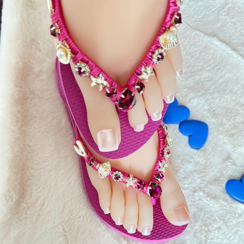 May include: Pink flip-flops with a jeweled strap featuring pink and gold accents. The strap is decorated with seashells, stars, and rhinestones.