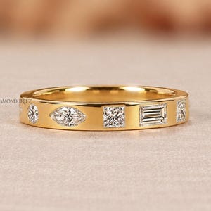 May include: A yellow gold band ring featuring a series of alternating diamond shapes, including round, marquise, princess, and baguette cuts set flush into the metal. The ring displays the text DIAMONDBREEZ on the left and bottom right.