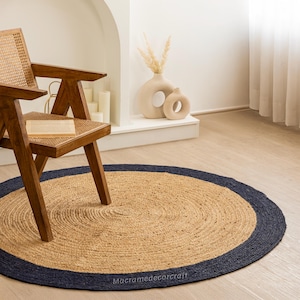 May include: A round jute rug with a navy blue border. The rug is placed on a light wood floor in a living room setting. A wooden chair with a woven seat sits on the rug.