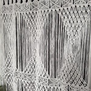 May include: White macrame wall hanging with a geometric pattern. The hanging is made of cotton cord and has a fringe at the bottom.