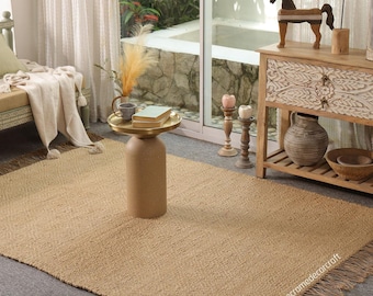 May include: A rectangular, woven jute rug with a fringed edge. The rug is a natural tan color and has a subtle geometric pattern. The rug is placed on a gray floor, and is part of a home decor setting.