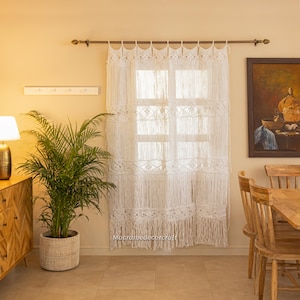 May include: White macrame curtain hanging in a room with a wooden table and chairs. The curtain is made of cotton cord and has a fringe at the bottom. The curtain is hung on a brass rod.