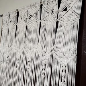 May include: White macrame wall hanging with a geometric pattern. The hanging is made of cotton cord and is attached to a metal rod.