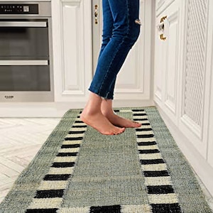 May include: A gray and black striped runner rug with a white border. The rug is made of natural fibers and has a textured surface. The rug is placed in a kitchen with white cabinets and a stainless steel oven.