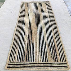 May include: A rectangular runner rug crafted from braided natural jute fibers. The design features a repeating pattern of vertical stripes in alternating shades of black, white, and tan, with horizontal striped borders at both ends of the rug.