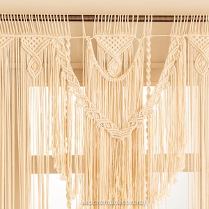 Puede incluir: Cortina de macramé color crema con intrincados patrones tejidos y un borde con flecos. La cortina está suspendida de una barra de madera y presenta diseños geométricos y un elemento central en forma de V. El texto "Macramedecorcraft" es visible en la parte inferior.