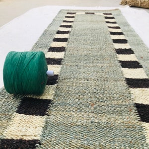 Pode incluir: Uma bola de fio verde está sobre um tapete tecido com uma borda xadrez preta e branca. O tapete é feito de fibras naturais e tem um visual rústico.