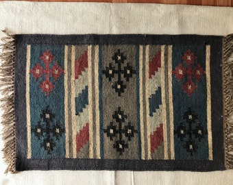 Puede incluir: Una alfombra rectangular con un patrón geométrico en tonos de azul, rojo, marrón y negro. La alfombra tiene flecos en los cuatro lados.
