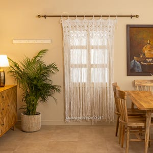 May include: White macrame curtain hanging in a window with a wooden dining table and chairs in the foreground. The curtain is made of cotton cord and has a fringe at the bottom. The curtain is hung on a brass rod.
