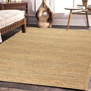 Puede incluir: Una alfombra de fibra natural con una textura tejida en color marrón claro. La alfombra tiene un sutil patrón a rayas y está hecha de yute.