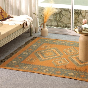 May include: An orange and teal geometric patterned rug with a fringe border. The rug features diamond and starburst designs. The text "Macramedecorcraft" is printed on the rug. The rug is on a gray floor.