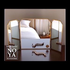 Peut inclure: Un miroir de coiffeuse à trois volets avec un cadre doré. Le miroir est ouvert, reflétant une scène de chambre. Deux rangées de bijoux perlés et une petite pièce de monnaie sont sur la surface devant le miroir. Le texte "NOYA box & terrarium" est visible.