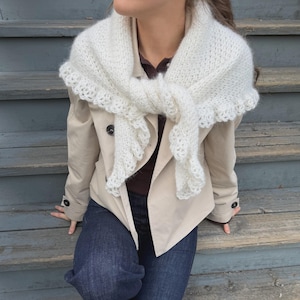 Può includere: Uno scialle triangolare in maglia bianca con un delicato bordo in pizzo smerlato, indossato sopra una giacca beige abbottonata, una camicia a colletto marrone scuro e jeans blu scuro. Lo scialle presenta una consistenza morbida e soffice, annodato sul davanti.