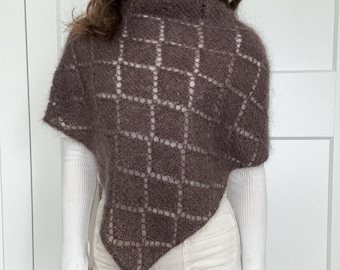 May include: A brown, open-weave poncho with a diamond pattern. The poncho is worn over a white turtleneck and white pants. The garment has a soft, textured appearance and a triangular shape.