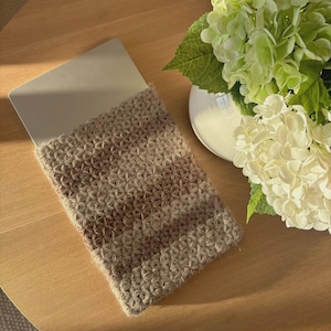 Op de afbeelding: Een laptophoes in een neutraal kleurenpalet, met een textuur, geweven ontwerp. De hoes ligt op een houten oppervlak, naast een vaas met witte en groene bloemen. De hoes lijkt gemaakt van een zachte, natuurlijke vezel.