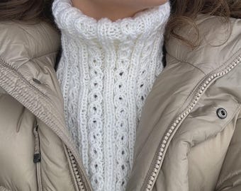 May include: A white, knitted cowl neck warmer with a textured pattern. The neck warmer is made of a soft, chunky yarn and is worn under a beige puffer jacket. The cowl neck is a winter accessory.