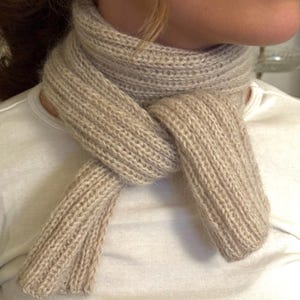 Può includere: Una sciarpa beige lavorata a maglia è annodata attorno al collo. La sciarpa è realizzata con un filato morbido e strutturato e presenta un motivo a coste. Le estremità della sciarpa pendono e la sciarpa è indossata sopra una maglietta bianca. La sciarpa è di colore neutro.