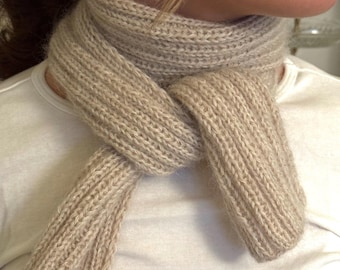 May include: A beige, knitted scarf is tied around the neck. The scarf is made of a soft, textured yarn and has a ribbed pattern. The ends of the scarf hang down, and the scarf is worn over a white shirt. The scarf is a neutral color.