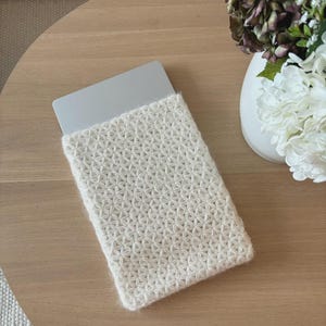 May include: A rectangular laptop sleeve featuring a cream-colored, textured crochet pattern with a star-like stitch design. The sleeve is constructed from soft yarn and partially encases a silver laptop, showing the top edge of the device.