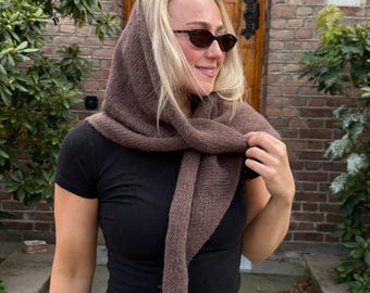 May include: A brown knitted hood and scarf set. The hood covers the head and neck, with the scarf portion draped around the shoulders and tied in front. The set is made of a soft, textured yarn, perfect for cooler weather.