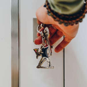 Puede incluir: Un llavero plateado con una figura que sostiene una flor y una llave adjunta.