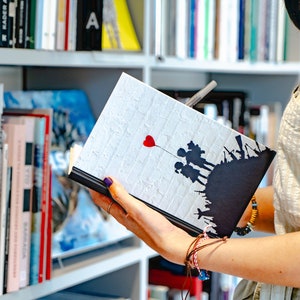 Könnte beinhalten: Ein weißes Notizbuch mit einer Schwarzweißillustration von Kindern, die einen roten Herzluftballon halten. Das Notizbuch hat einen Hintergrund mit Ziegelmuster.