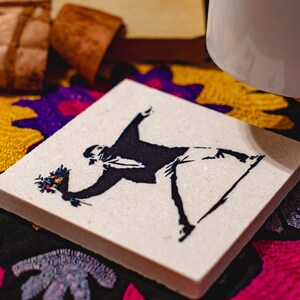 May include: A white square coaster with a black and white silhouette of a person throwing a bouquet of flowers. The coaster is on a colorful fabric background.