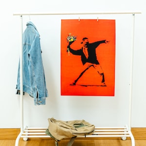 May include: A white clothes rack with a denim jacket hanging on a hanger, a brown rucksack on the bottom shelf, and a red poster with a black silhouette of a person throwing flowers.